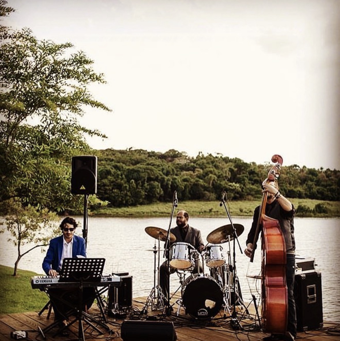 Trio de piano, bateria e contrabaixo tocando à beira do lago